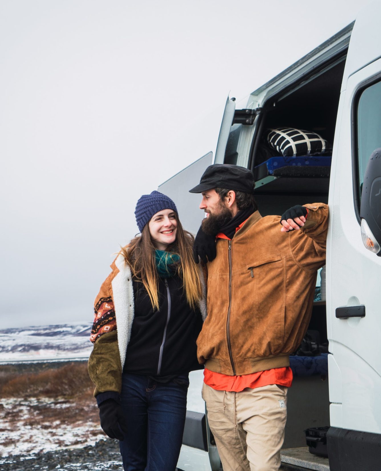 Content happy couple in outwear looking at each other standing near van in snowy valley of Iceland.
